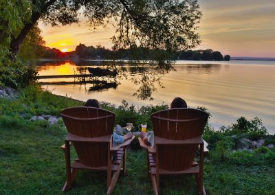 A couple relaxing with drinks sitting in Muskoka chairs watching the sunset over the water